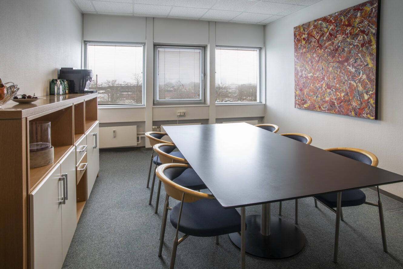 Lille mødelokale med et sort rektangulært bord, seks stole, et sideskab med forfriskninger, tre vinduer og et stort abstrakt maleri på væggen.