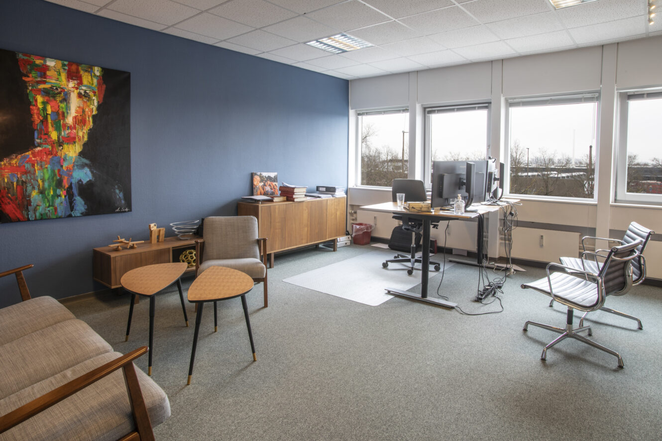 Moderne kontor med blå accentvæg, abstrakte portrætkunstværker, træmøbler, et skrivebord med computerskærme og flere stole, alt sammen oplyst af naturligt lys fra store vinduer.