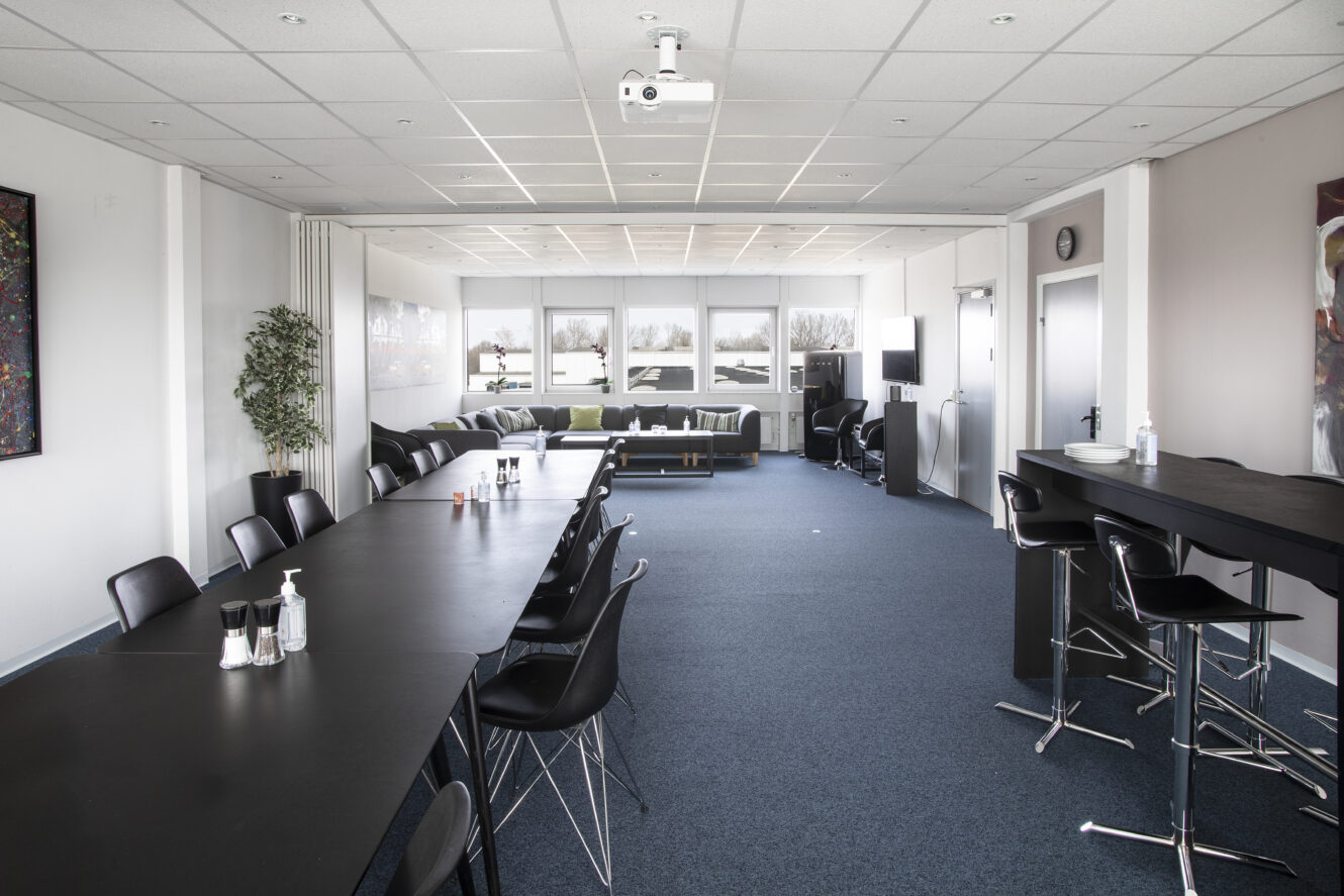 Et moderne konferencelokale med et langt sort bord, sorte stole, en projektor, et barområde og en lounge med sofaer ved store vinduer.