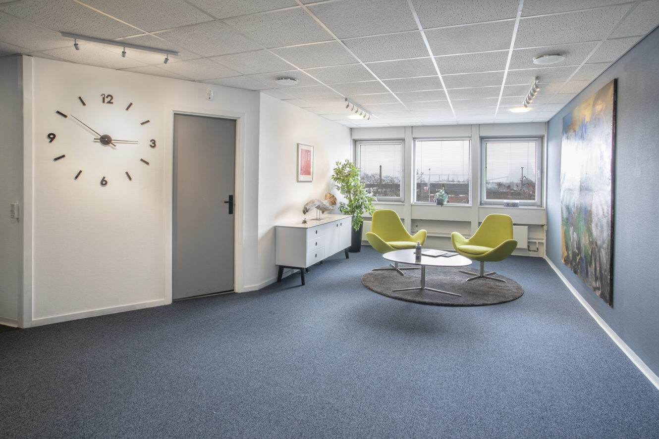 Moderne venteområde på kontoret med to grønne stole og et lille bord på et rundt tæppe, stort vægur, kunstværker og vinduer, der lukker naturligt lys ind.