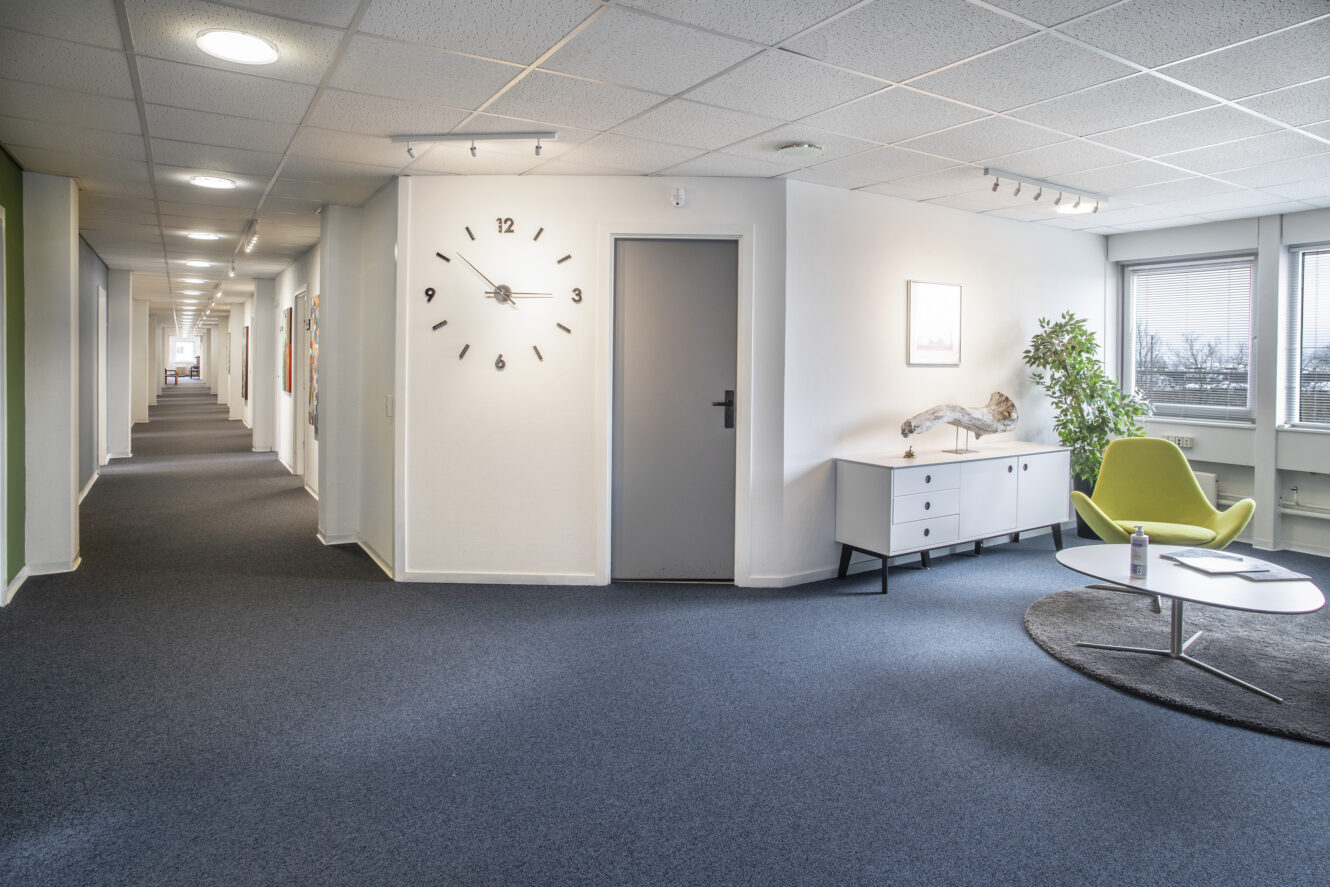 En moderne kontorgang med blåt gulvtæppe, et vægur over en grå dør, en grøn stol ved et rundt bord, en skænk og store vinduer, der lukker naturligt lys ind.