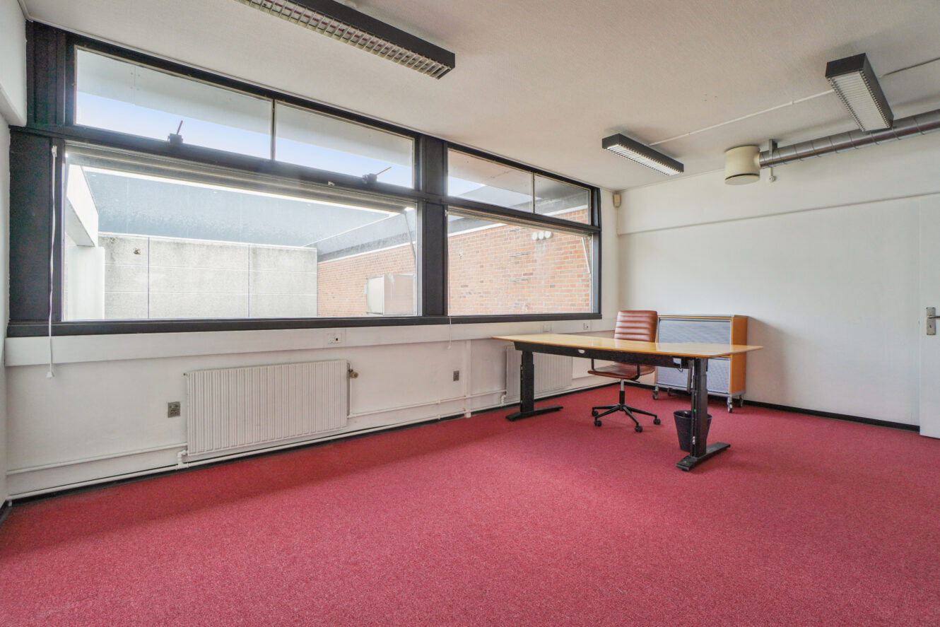 Et tomt kontor med rødt gulvtæppe, et stort vindue, hvide vægge, et træskrivebord, en kontorstol og en radiator under vinduet.