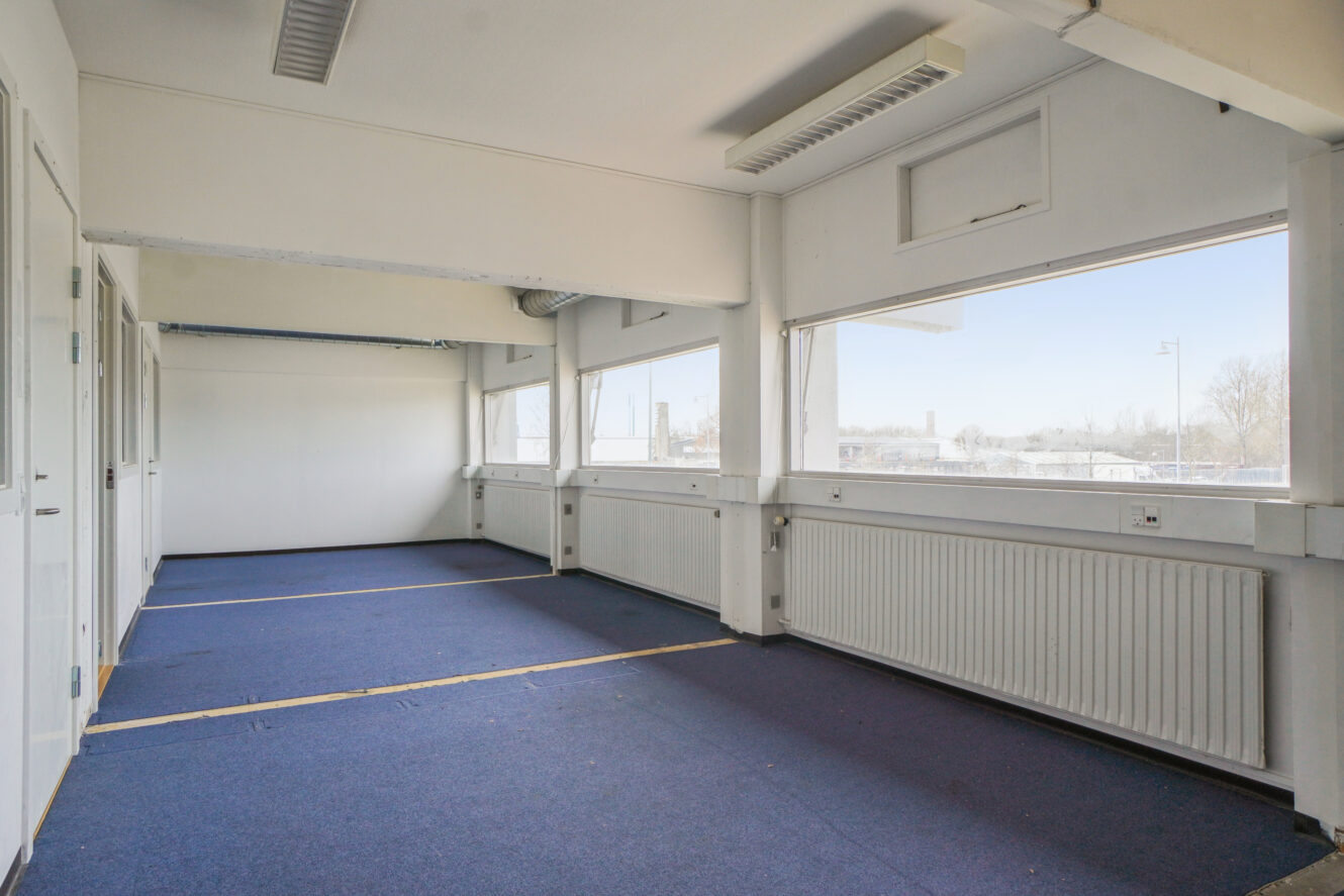 Et tomt rum med blåt gulvtæppe, hvide vægge, store vinduer, lysstofrør i loftet og radiatorer under vinduerne.