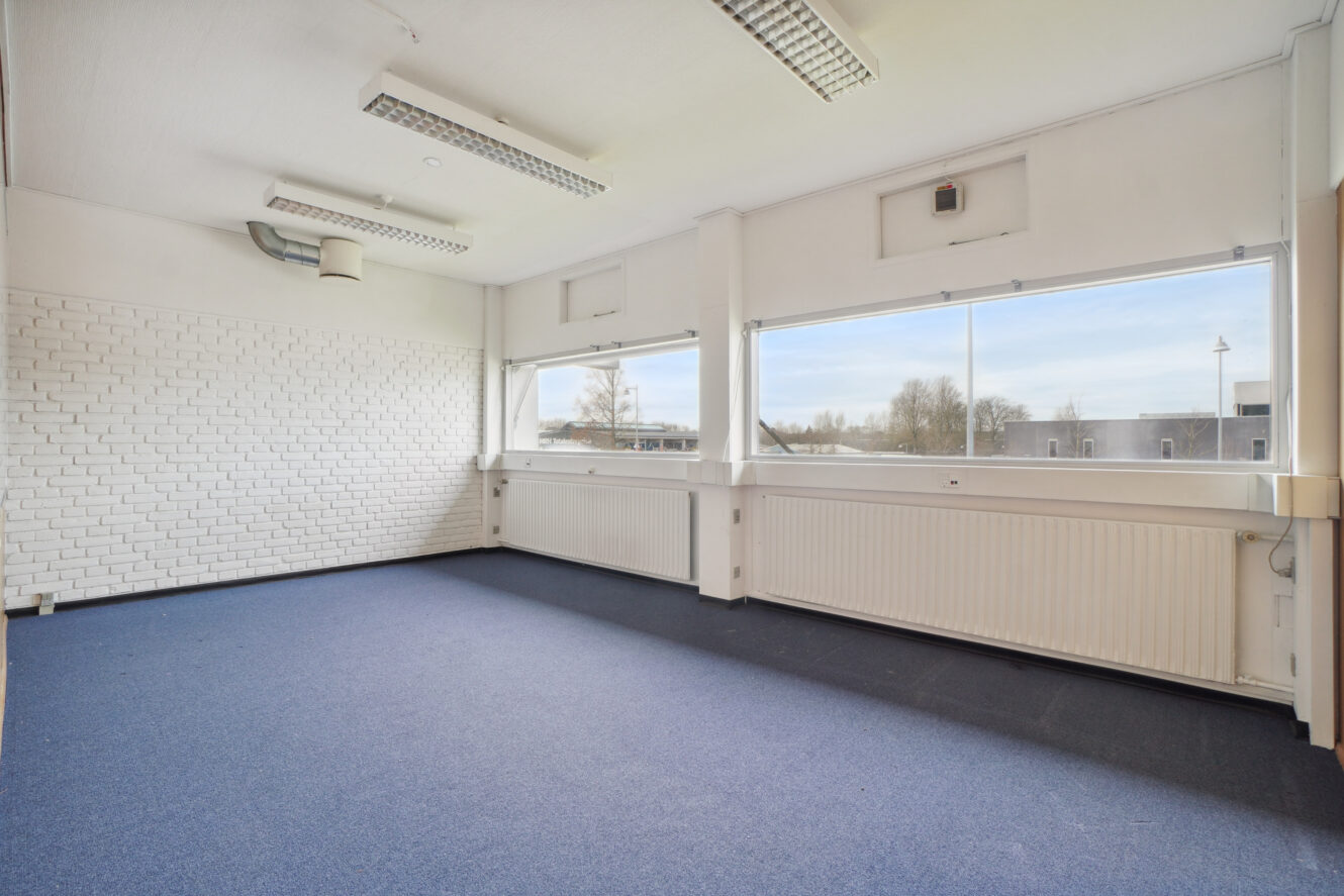 Tomt kontorlokale med hvide vægge, store vinduer, lysstofrør i loftet, blåt gulvtæppe og synlige radiatorer under vinduerne.