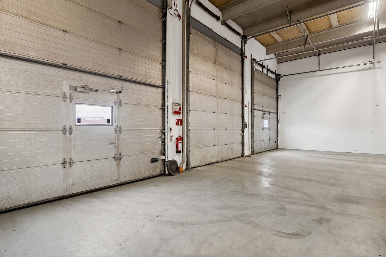 Tom industrihal med cementgulv, tre store porte med vinduer, en brandslukker monteret på væggen samt højt til loftet med synlige bjælker og belysning.
