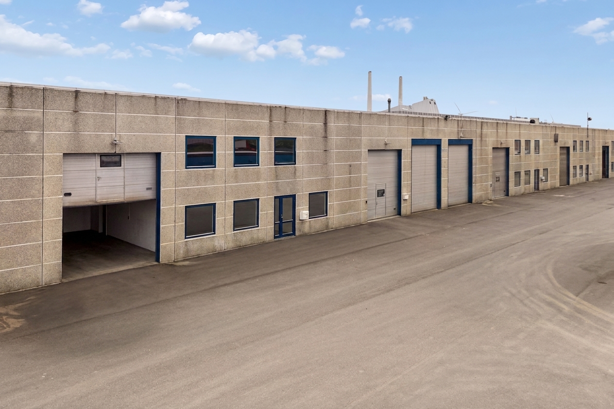 En række industrielle lagerbygninger med store garageporte og vinduer vender ud mod en asfalteret grund under en blå himmel med skyer.