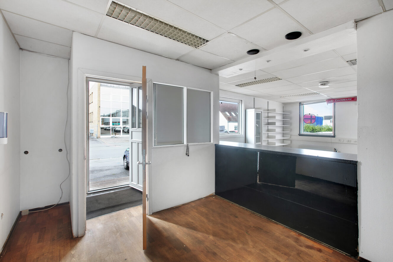 Et tomt kontorlokale med hvide vægge, trægulv, sort skranke, åbne vinduer og en dør, der fører ud til en gade med parkerede biler, der kan ses gennem glasset.