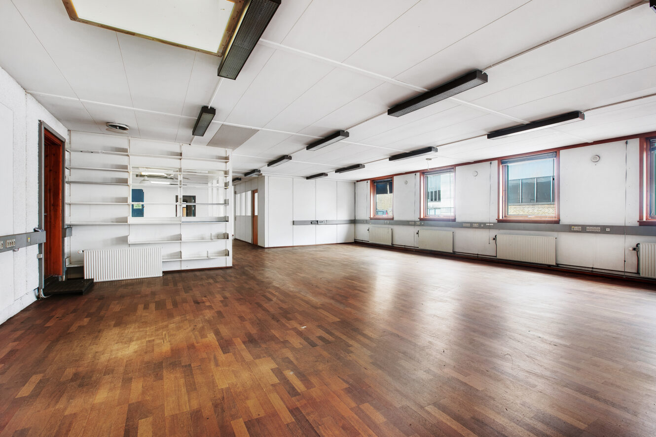 Rummeligt, tomt rum med trægulv, hvide vægge, store vinduer, indbyggede hylder og loftslamper.