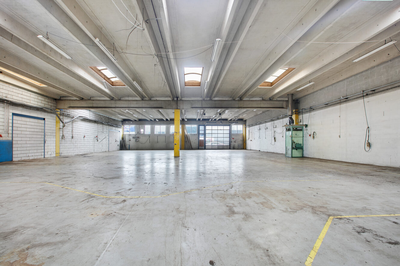 Stort tomt industrilager med betongulv, synlige loftsbjælker, store vinduer og fluorescerende belysning.