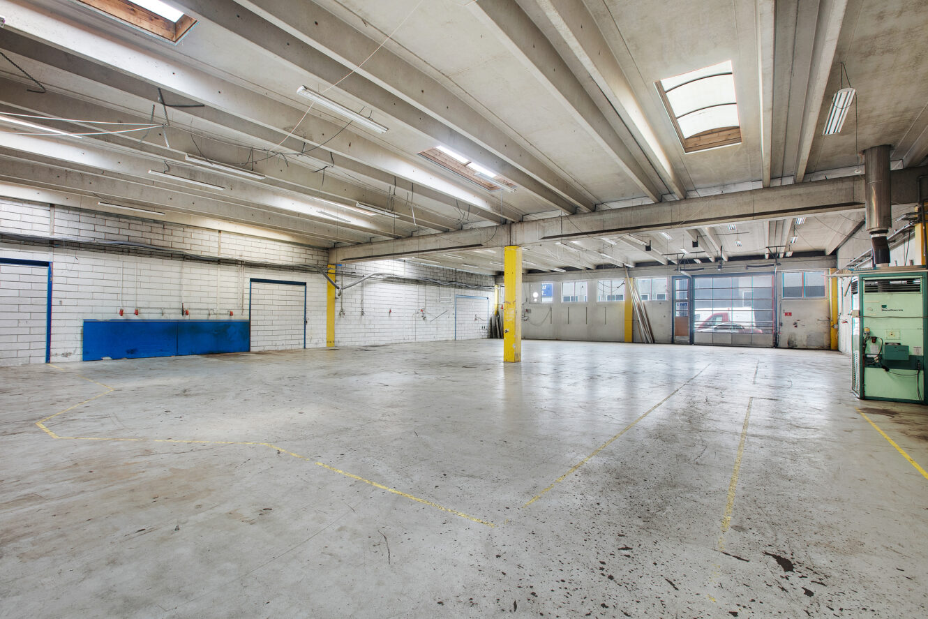 Stort, tomt industrilager med betongulv, synlige loftsbjælker, store vinduer og en gul støttesøjle.