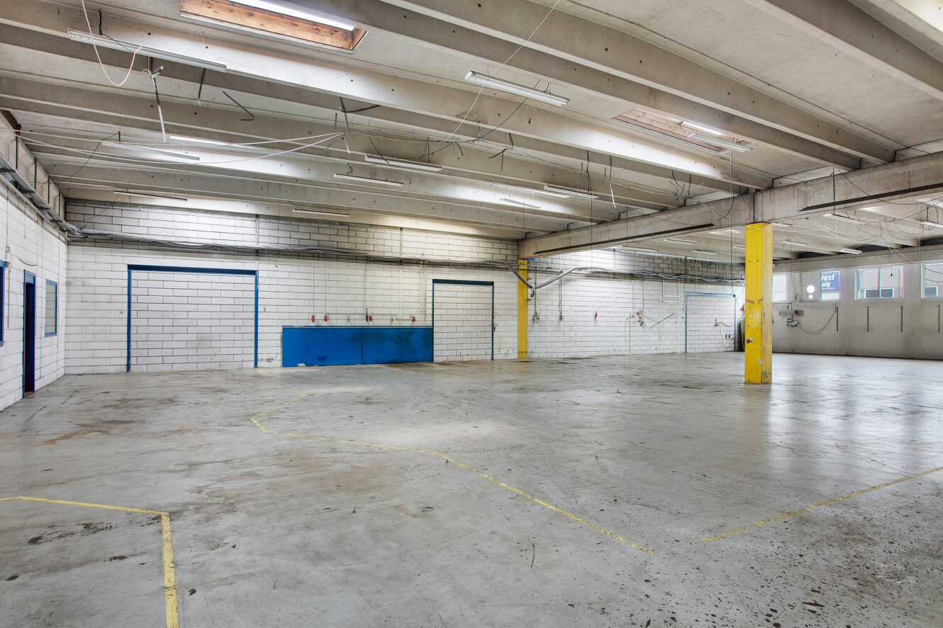 Et tomt industrilager med hvide murstensvægge, betongulv, synlige loftsbjælker og gule sikkerhedsmærker på jorden.