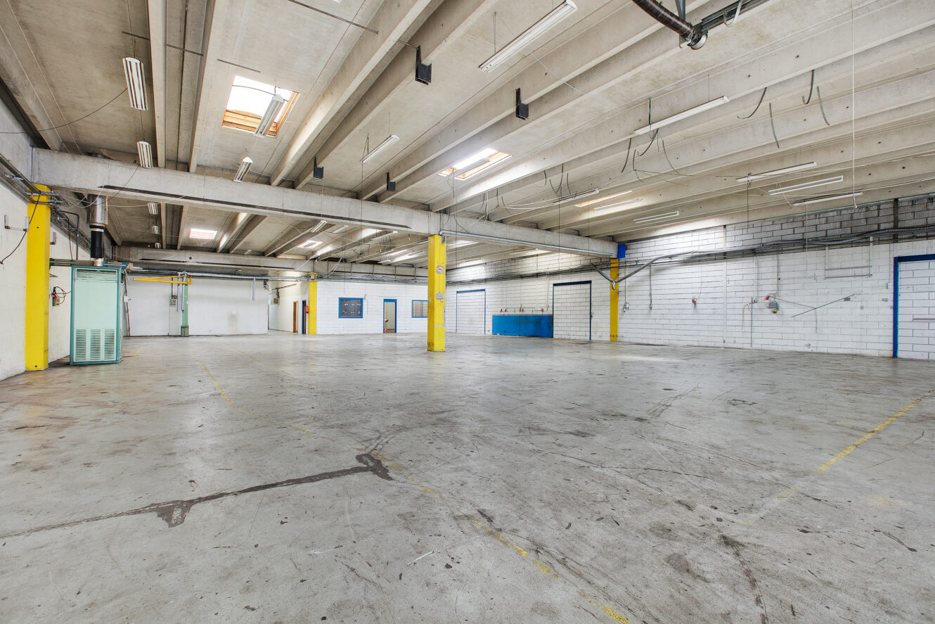 Stort, tomt industrilager med betongulv, højt til loftet, synlige bjælker, gule søjler og fluorescerende belysning.