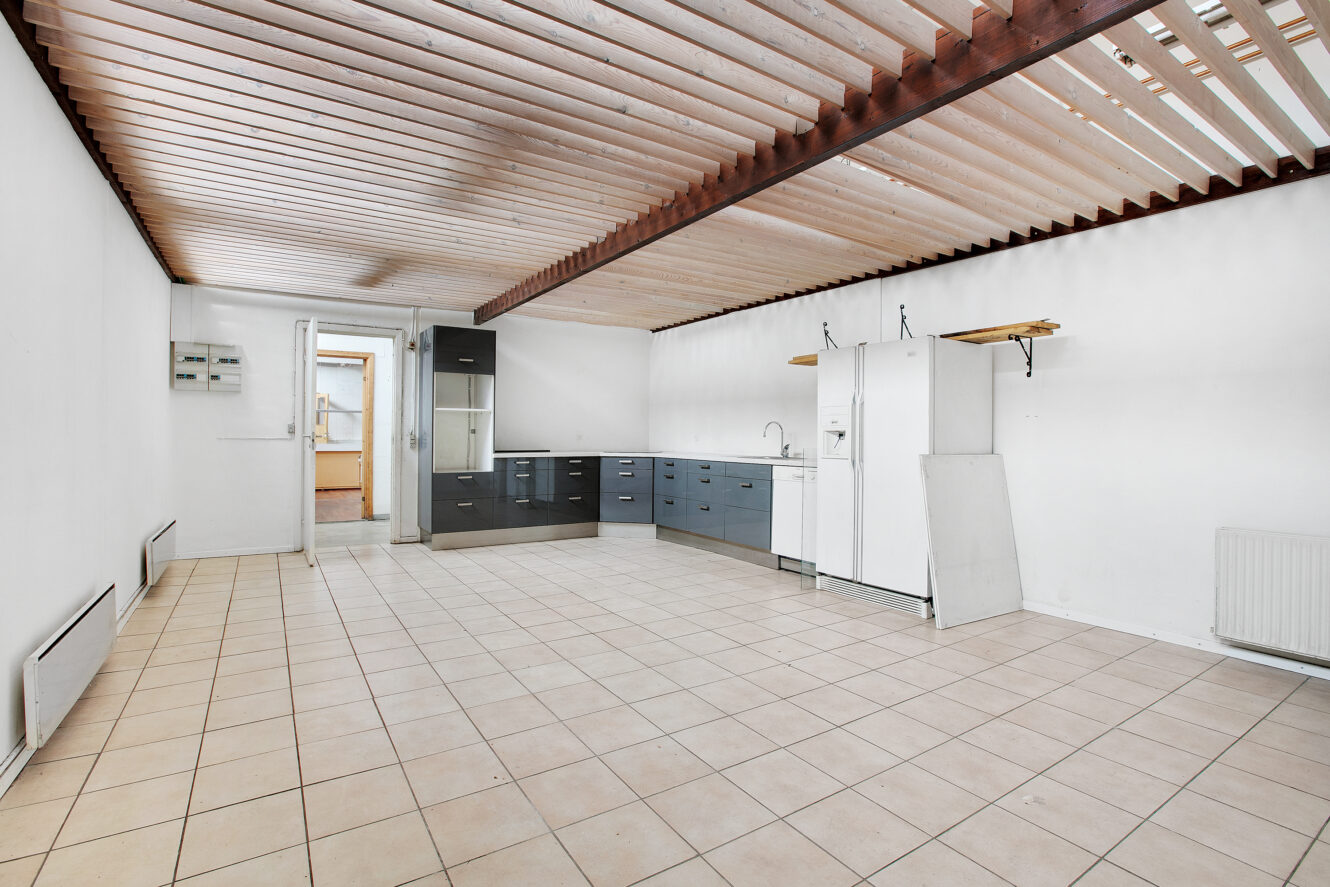 Rummeligt, umøbleret værelse med klinkegulv, hvide vægge, synlige loftslameller og et køkkenhjørne med skabe, køleskab og vask.