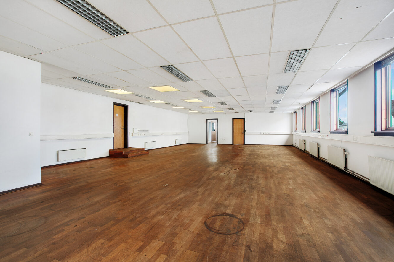 Et stort, tomt kontorlokale med trægulv, hvide vægge, loftslamper, flere døre og vinduer langs den ene væg.
