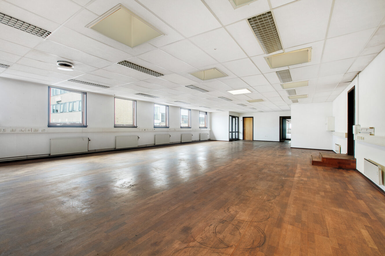 Stort, tomt kontorlokale med trægulv, hvide loftsplader, lysstofrør og flere vinduer langs den ene væg.