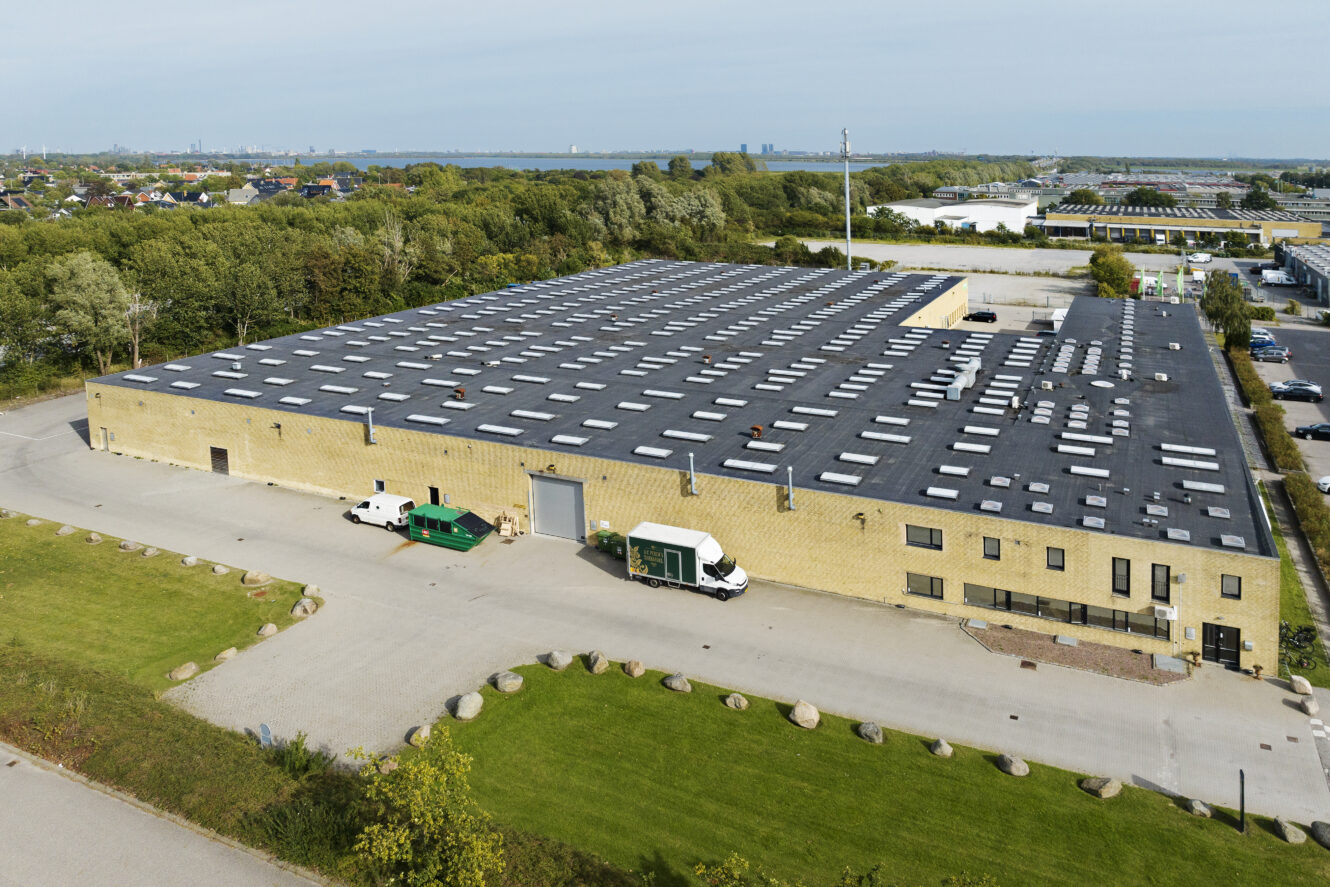 En stor industrihal med fladt tag og ovenlys er omgivet af træer og parkerede biler, set fra luften på en klar dag.