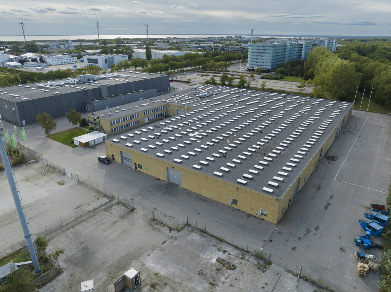 Luftfoto af et stort industrilager med fladt tag og mange ovenlysvinduer, omgivet af industribygninger, træer og vindmøller i baggrunden.