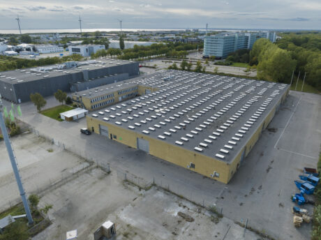 Luftfoto af et stort industrilager med fladt tag og mange ovenlysvinduer, omgivet af industribygninger, træer og vindmøller i baggrunden.