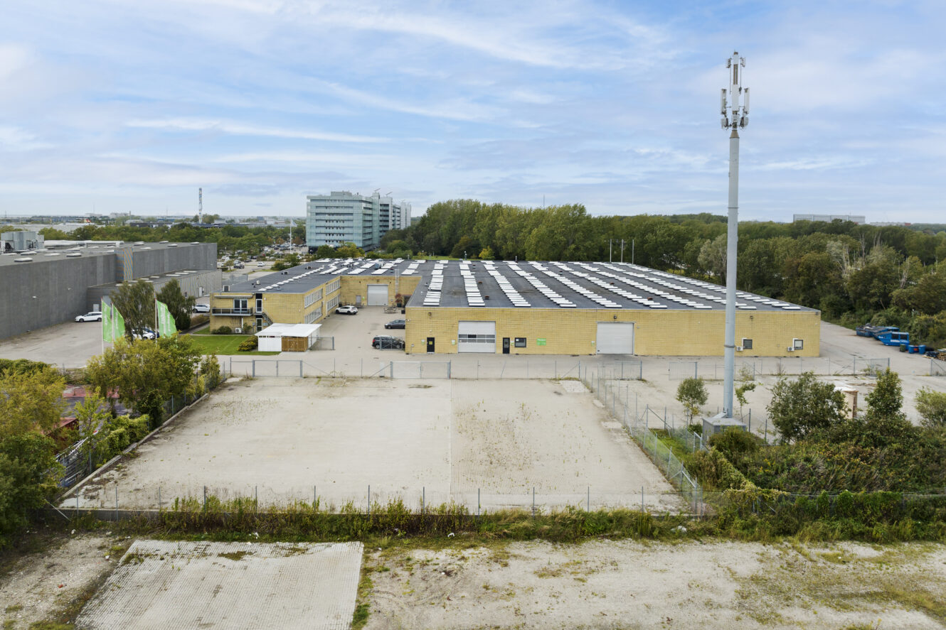 En stor industribygning med et fladt tag dækket af solpaneler, omgivet af træer, veje og andre kommercielle strukturer under en delvis overskyet himmel.