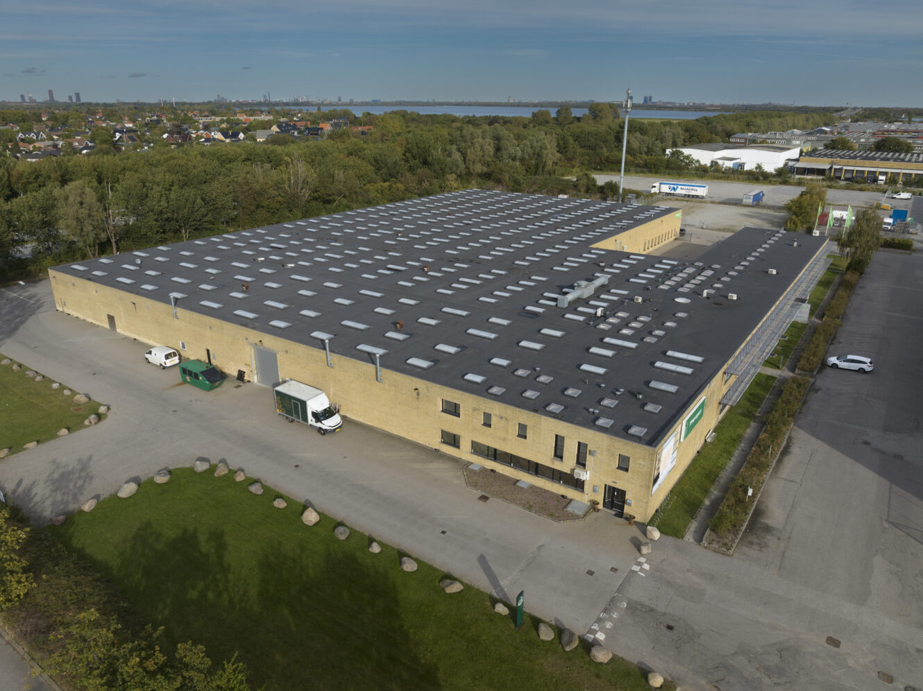 Luftfoto af et stort, rektangulært industrilager med fladt tag og læsseramper, omgivet af parkeringspladser og grønne områder.
