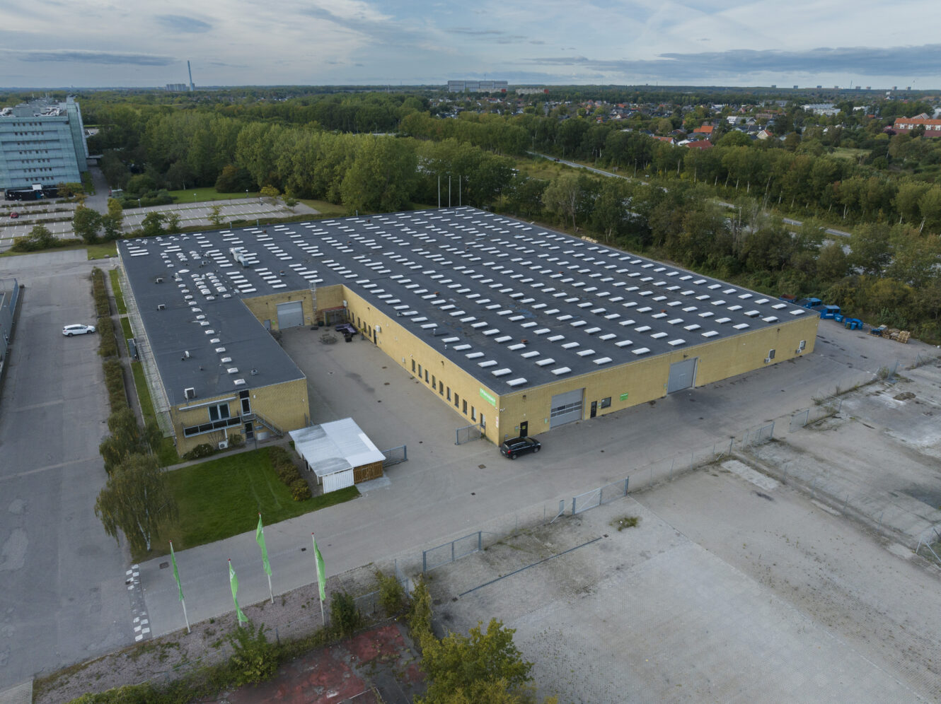 Luftfoto af et stort industrilager med fladt tag og flere ovenlysvinduer, omgivet af træer og bygninger i nærheden.