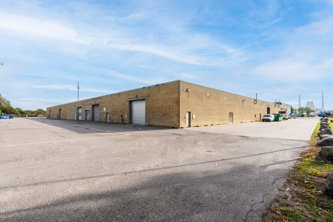 En stor, rektangulær lagerbygning i brun mursten med et par garageporte og et minimum af vinduer, der støder op til en tom parkeringsplads under en delvis overskyet himmel.