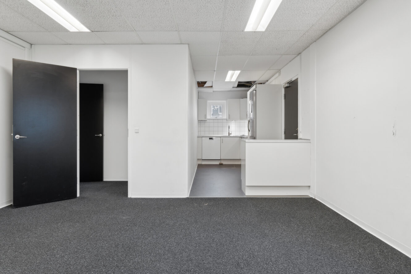 Et tomt kontorlokale med mørkt gulvtæppe, hvide vægge, en åben dør og et synligt tekøkken i baggrunden. Lysstofrør i loftet er tændt.