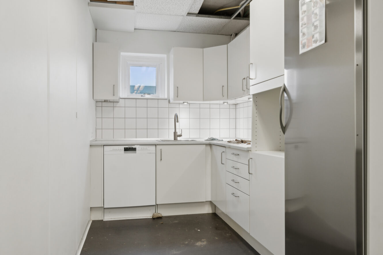 Lille, moderne køkken med hvide skabe, køleskab i rustfrit stål, opvaskemaskine og et enkelt vindue over vasken; synlige loftsplader.