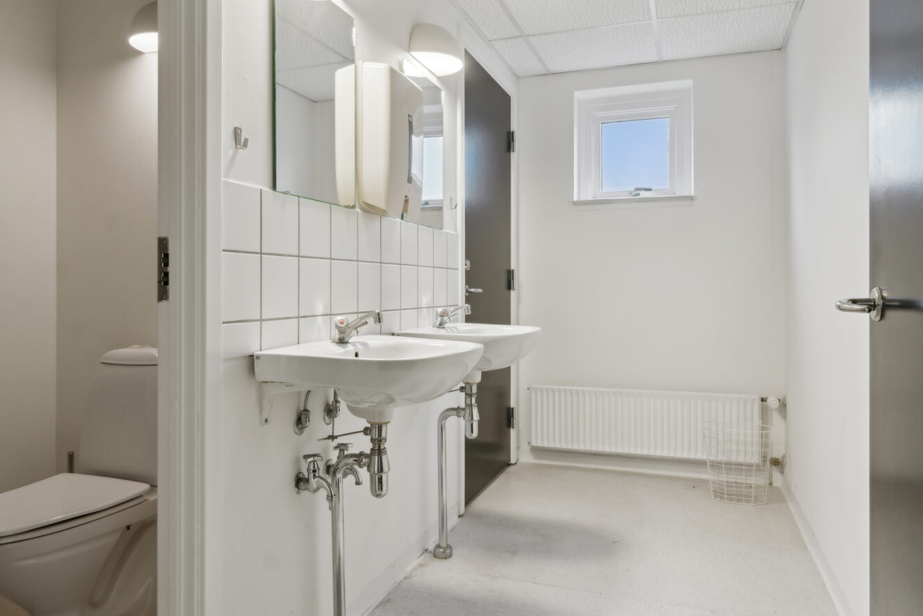 Et rent, moderne badeværelse med to hvide håndvaske, et vægspejl, et toilet, et lille vindue og en radiator under vinduet.