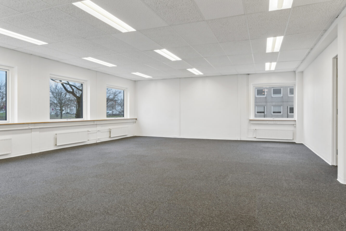 Tomt kontorlokale med gråt gulvtæppe, hvide vægge, store vinduer og lysstofrør i loftet.