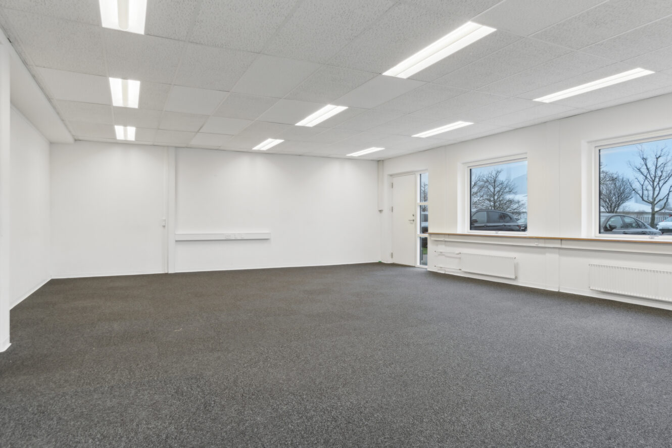 Tomt kontorlokale med gråt gulvtæppe, hvide vægge, nedhængt loft med lysstofrør og tre vinduer, der viser parkerede biler og træer udenfor.