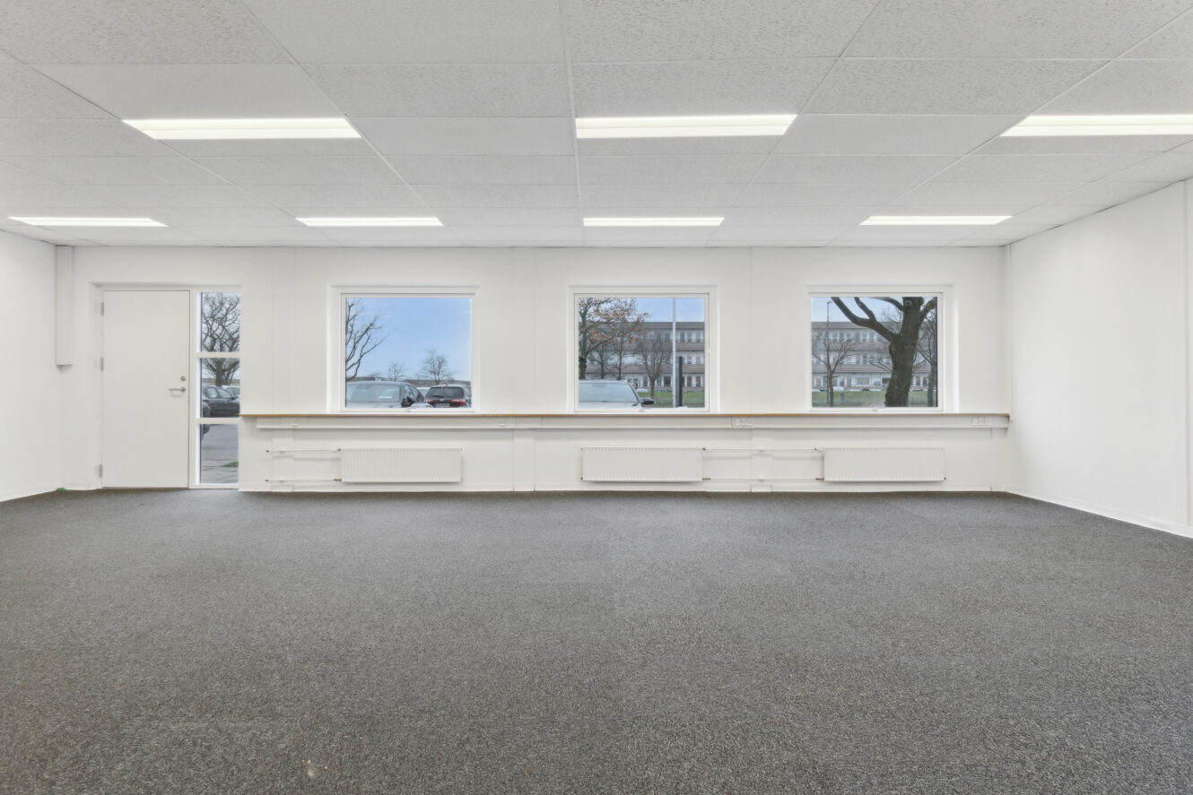 Tomt rum med gråt gulvtæppe, hvide vægge, tre store vinduer, lysstofrør i loftet og en lukket dør til venstre. Træer og bygninger er synlige uden for vinduerne.