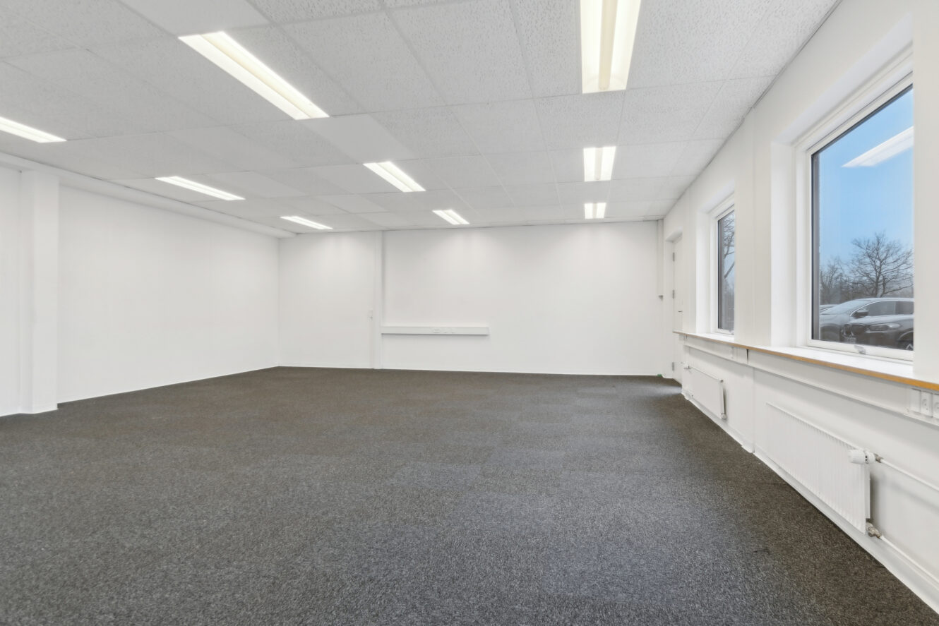 Tomt kontorlokale med gråt gulvtæppe, hvide vægge, nedhængt loft med lysstofrør og store vinduer, der viser en parkeret bil udenfor.