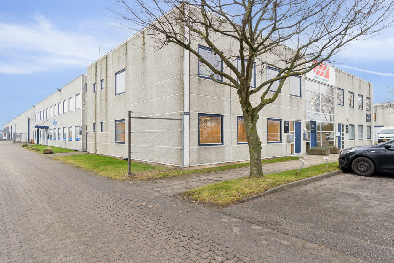 En toetagers industriel kontorbygning med store vinduer, gråt ydre og et træ foran, beliggende på en asfalteret gade med parkerede biler.