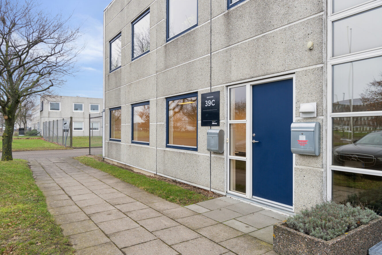 En moderne toetagers betonbygning med blå døre og vinduesrammer, mærket 39C, ved siden af en asfalteret gangsti og et græsareal med træer.