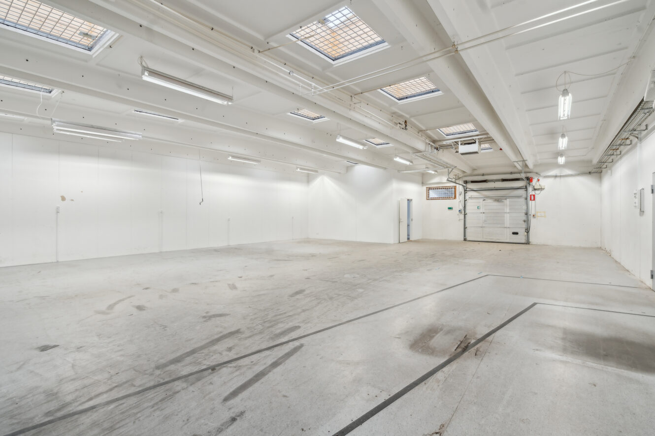 Stort tomt industrilager med hvide vægge, højt til loftet, flere ovenlysvinduer, lysstofrør og en lukket rulleport på bagsiden.