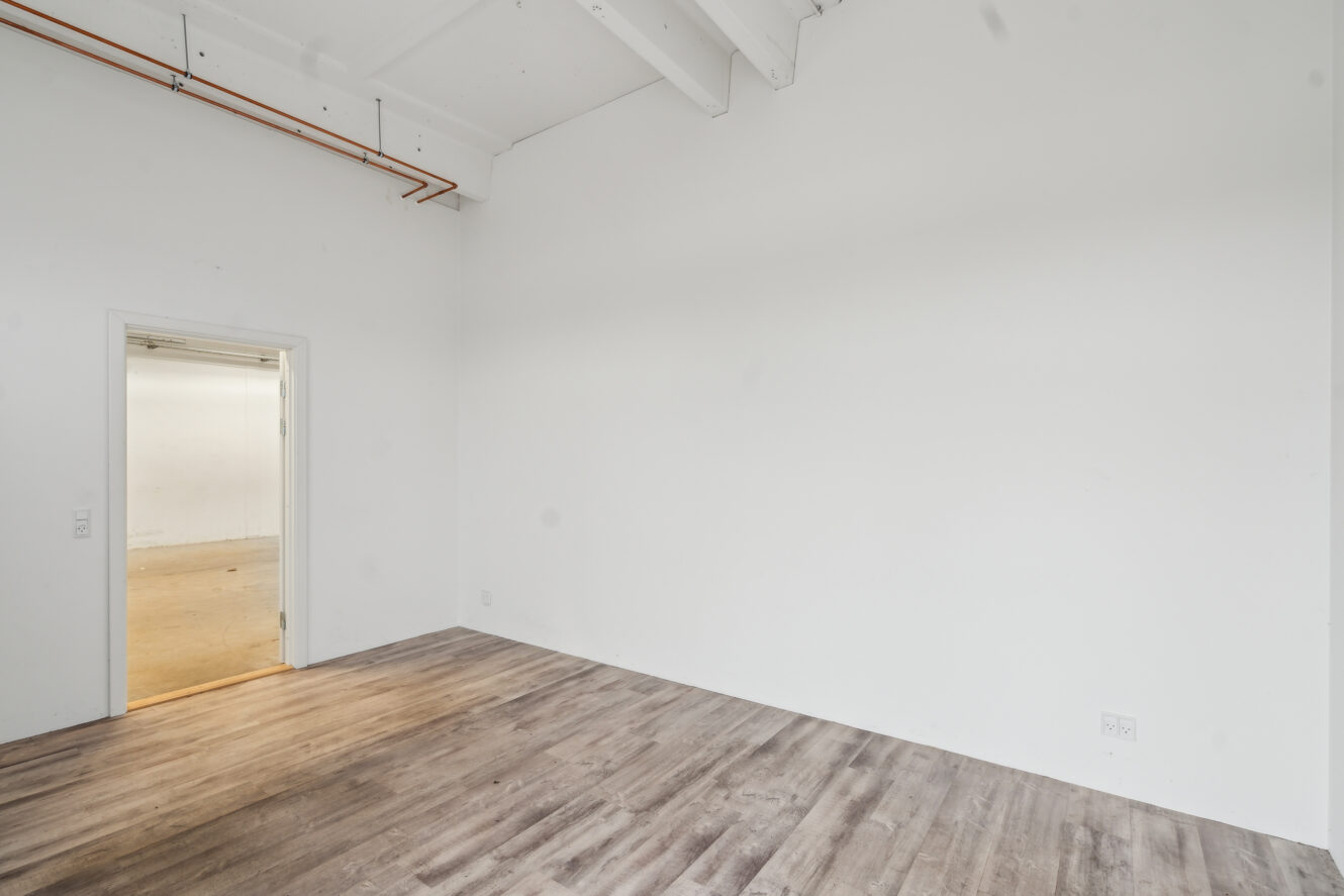 Tomt rum med hvide vægge, højt til loftet med synlige rør, trægulv og en åben døråbning, der fører til et andet rum med lyst gulv.