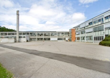 En stor, tom, asfalteret gårdsplads er omgivet af fleretagers bygninger i industristil med mange vinduer og en høj skorsten til venstre.