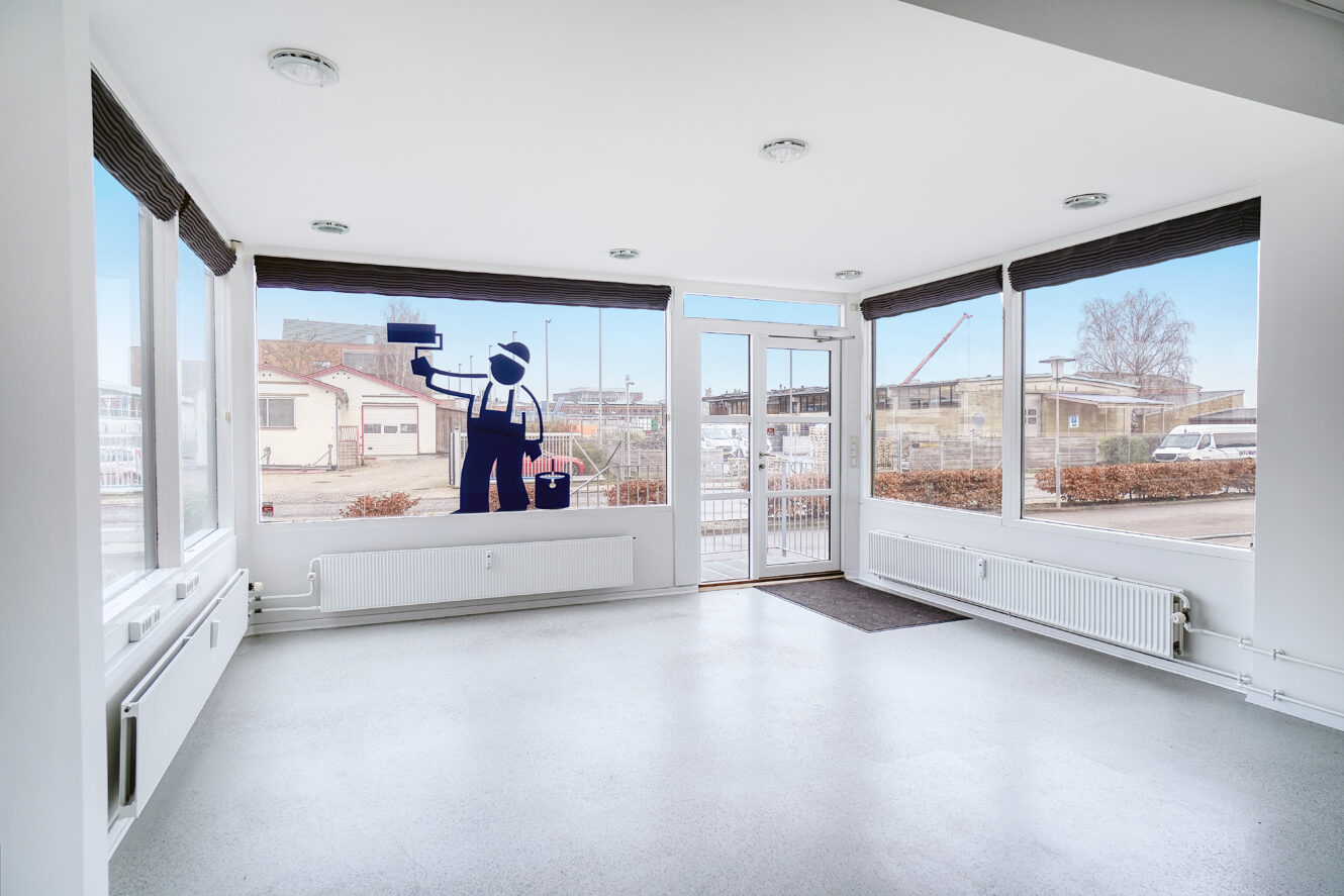 Tomt, lyst rum med store vinduer og en dør med en malersilhuet, der holder en rulle og en spand på det ene vindue. Udenfor kan man se entreprenørmaskiner.