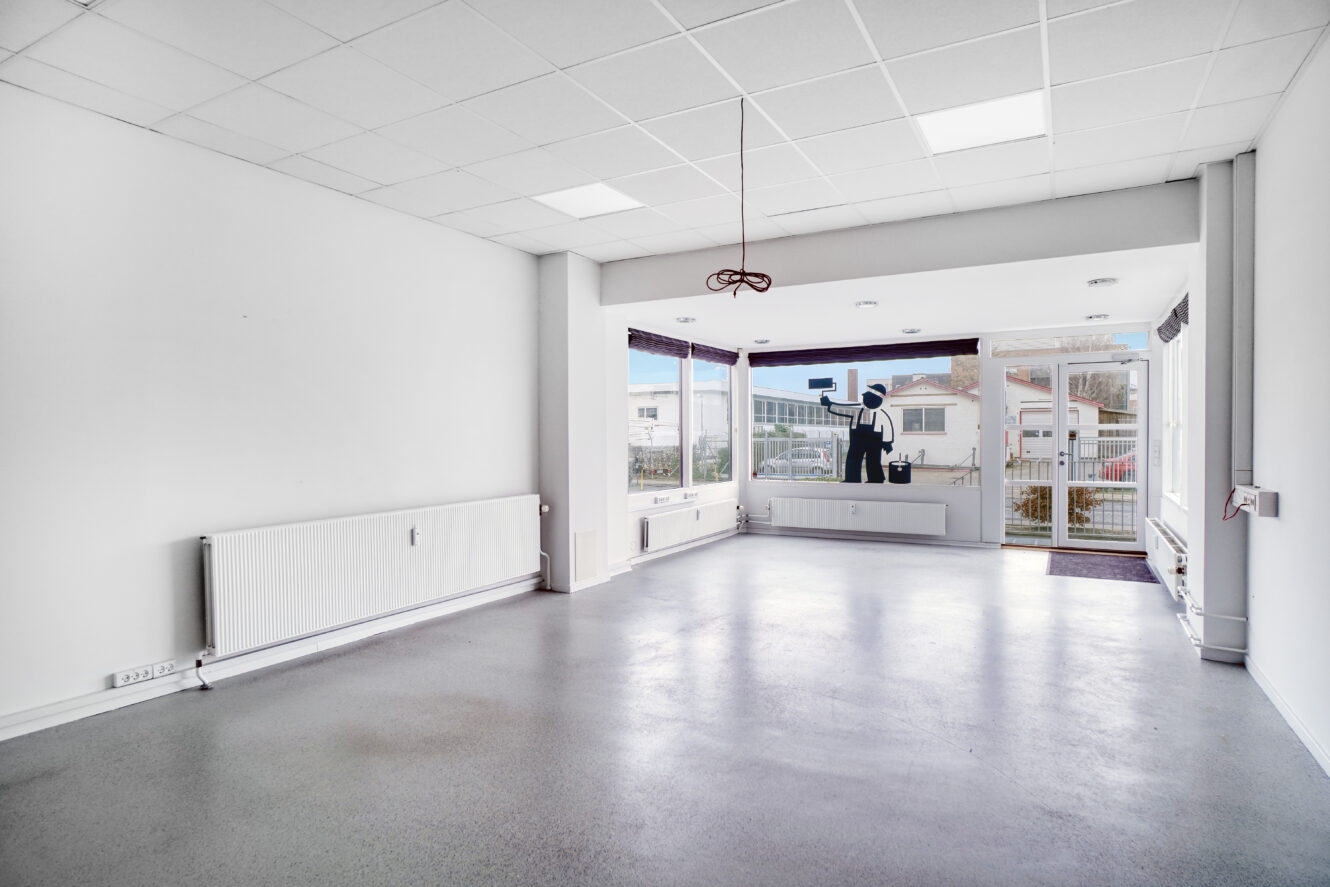 Tomt, umøbleret rum med hvide vægge, store vinduer, en radiator og en dør, der fører udenfor; man kan se gaden og bygninger gennem vinduerne.