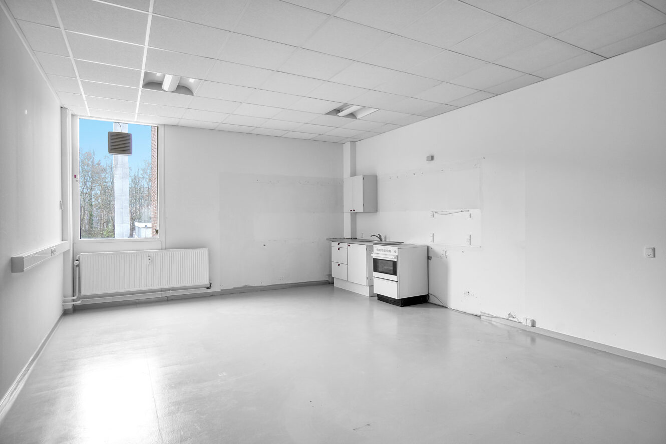 Tomt, umøbleret rum med hvide vægge, et lille tekøkken, et komfur, en radiator under et vindue og stærkt naturligt lys, der kommer ind fra venstre side.