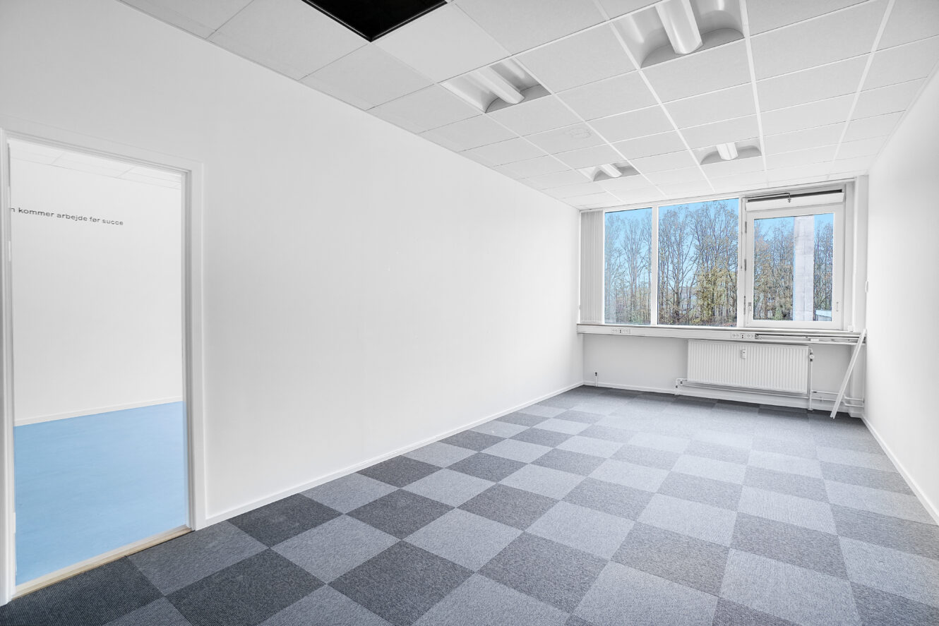 Et tomt kontorlokale med gråternet tæppe, hvide vægge og store vinduer med udsigt til træer; en åben dør fører ind til et rum med blåt gulv.