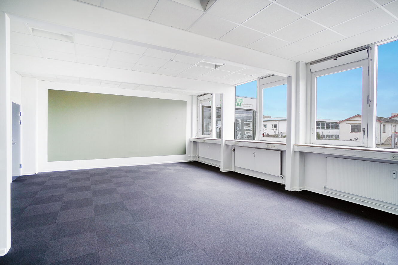 Tomt kontorlokale med lyse vægge, store vinduer, gråt ternet tæppe og en grøn accentvæg. Naturligt lys kommer ind gennem vinduerne og viser bygningerne udenfor.