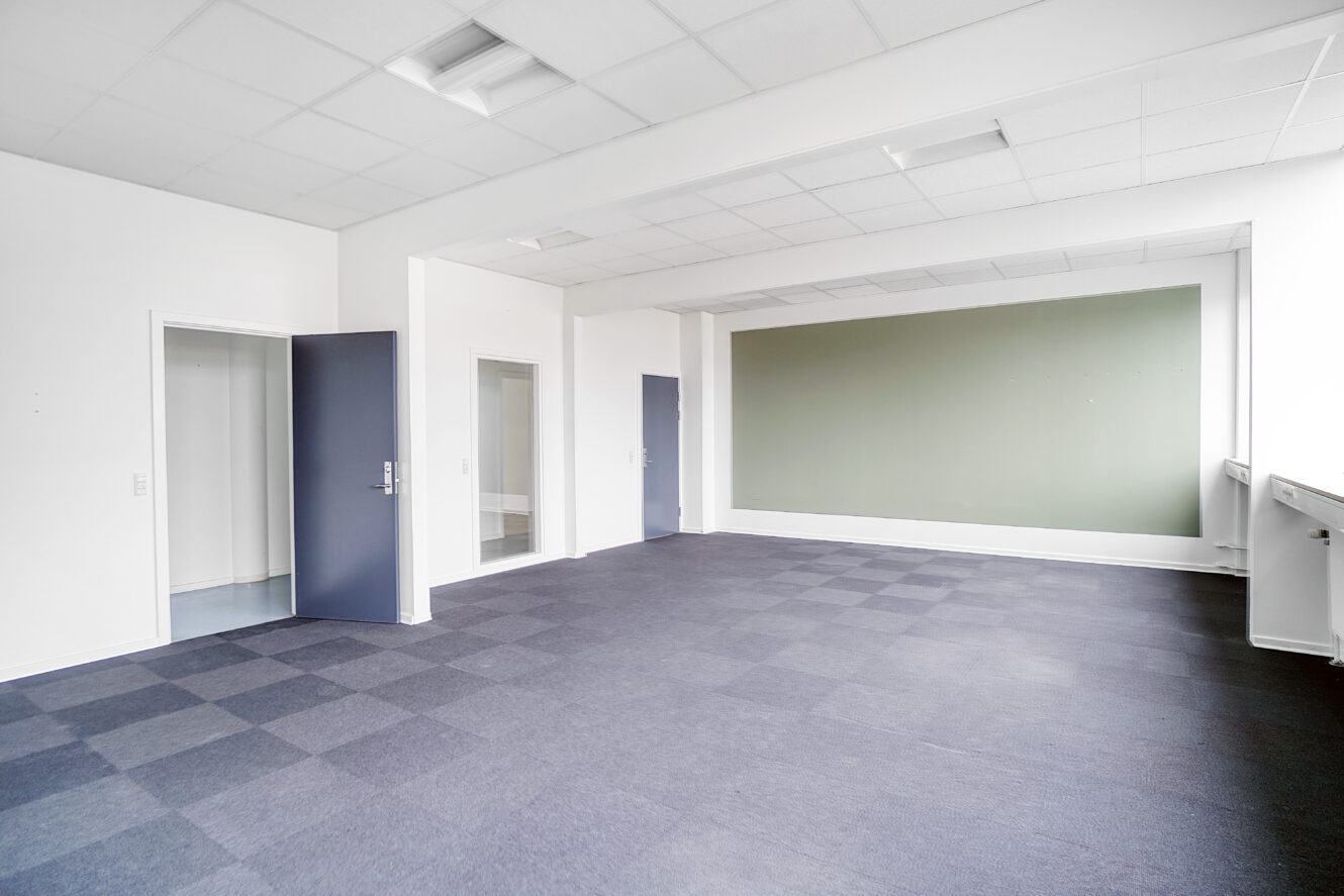 Tomt rum med grå tæppefliser, hvide vægge, en grøn accentvæg og lysstofrør i loftet. Der er to døre og flere vinduer langs den ene væg.