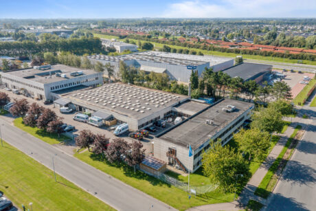 Luftfoto af et industrikompleks med flere bygninger, lastbiler og parkeringspladser, omgivet af træer og veje i et forstadsmiljø.