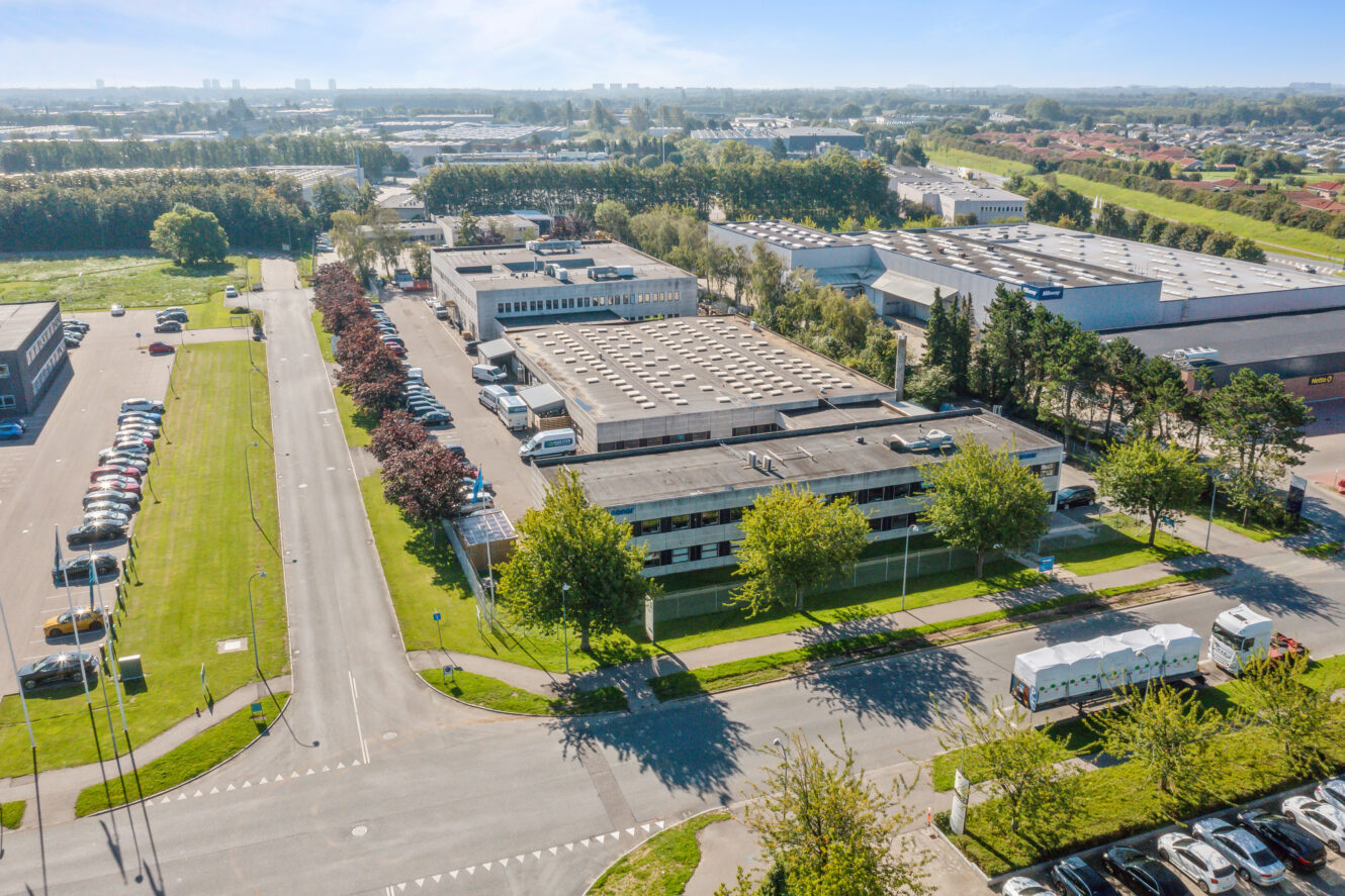 Luftfoto af en industripark med flere store lagerbygninger, parkeringspladser, træer og omkringliggende veje på en klar, solrig dag.