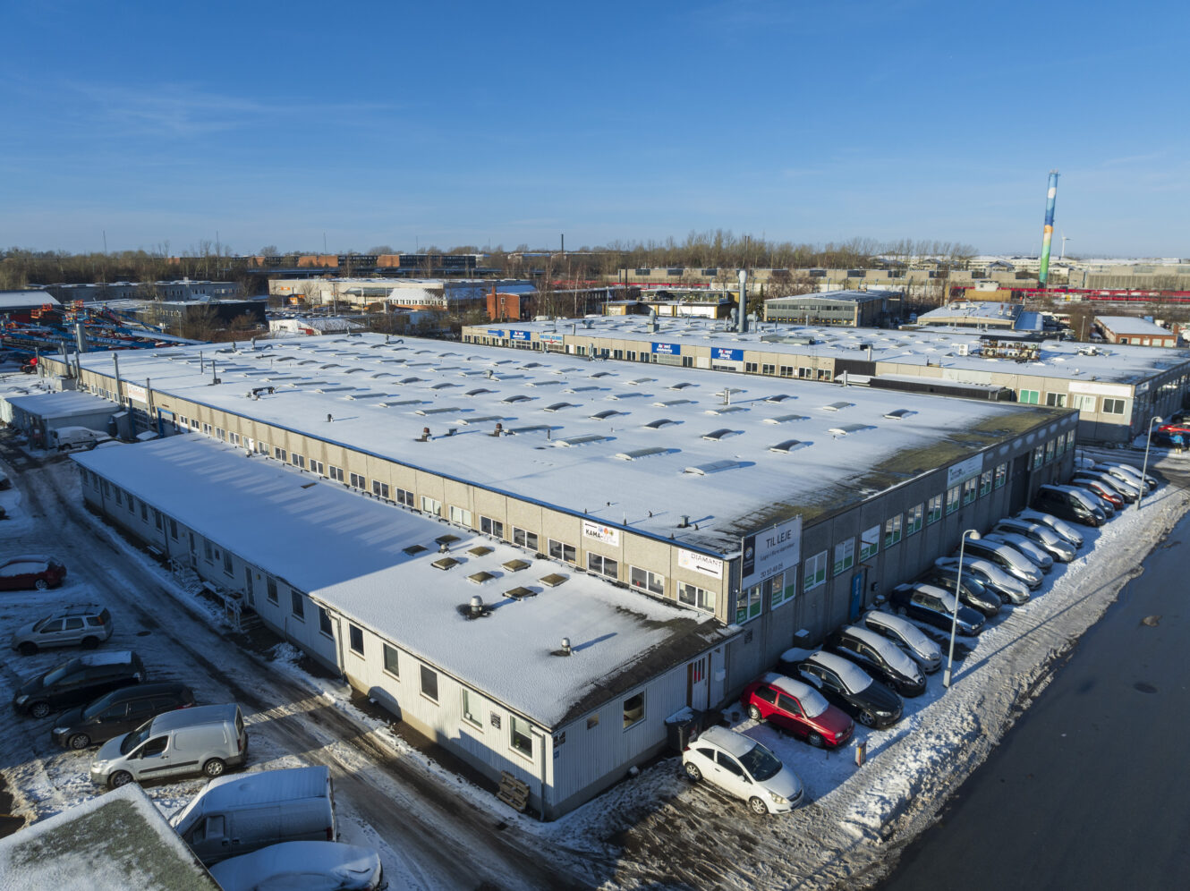 En stor industribygning med et snedækket tag, omgivet af parkerede biler og delvist snedækket jord under en klar blå himmel.