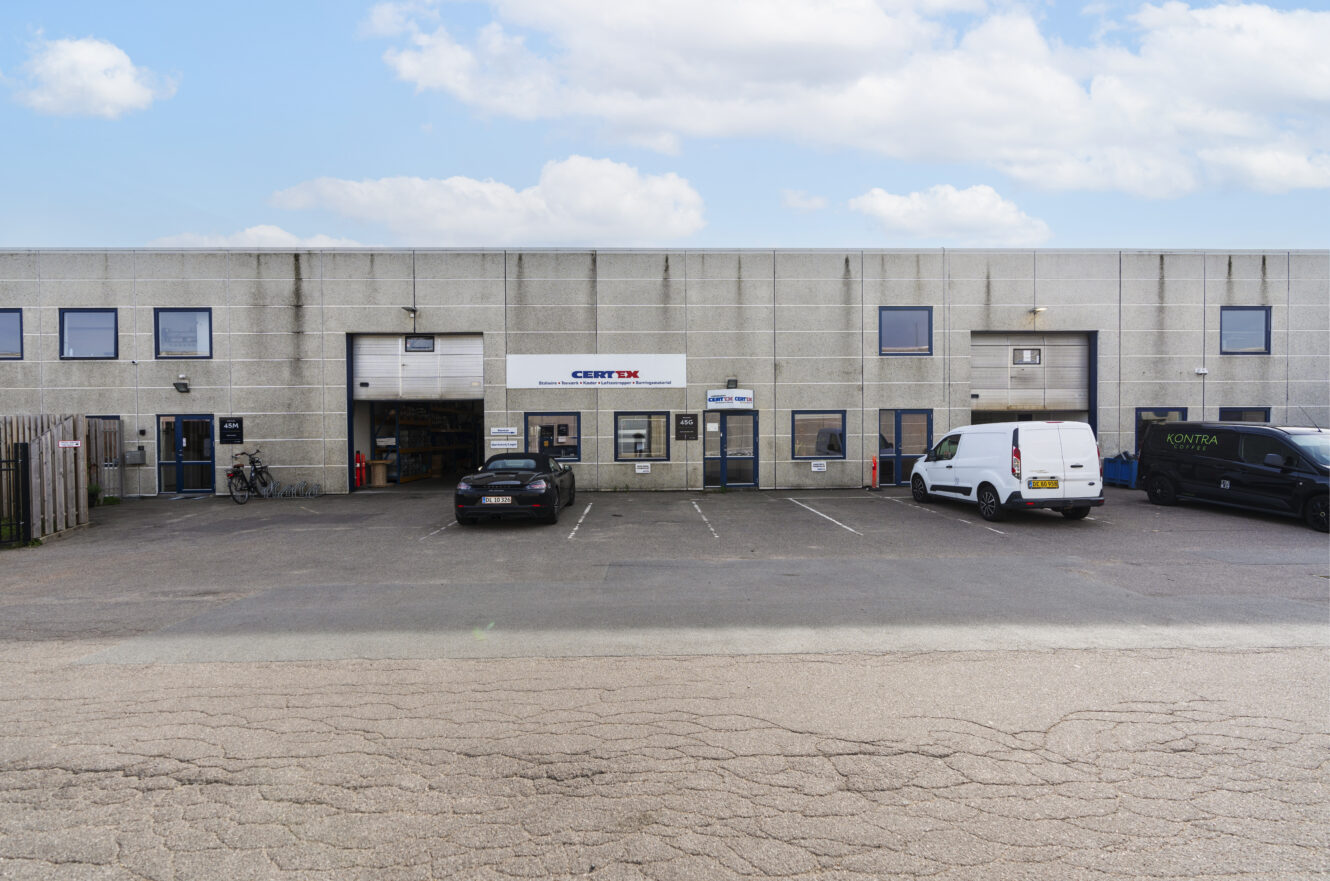 En grå industribygning med flere indgange, to parkerede varevogne, en bil og cykler foran under en delvist overskyet himmel.