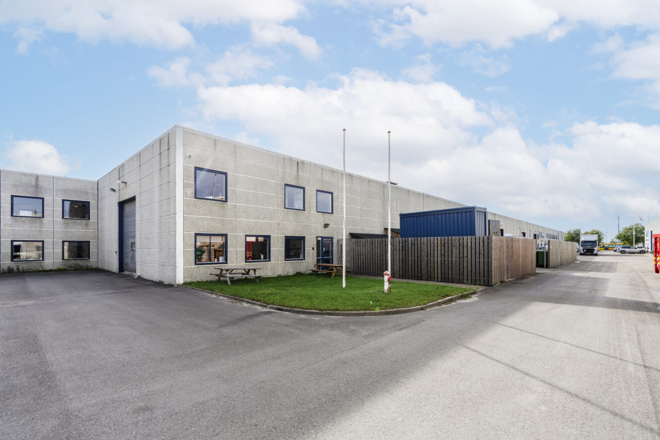 En stor industribygning med flere vinduer, en garageport, et indhegnet område og et picnicbord på en lille græsplæne under en delvis overskyet himmel.