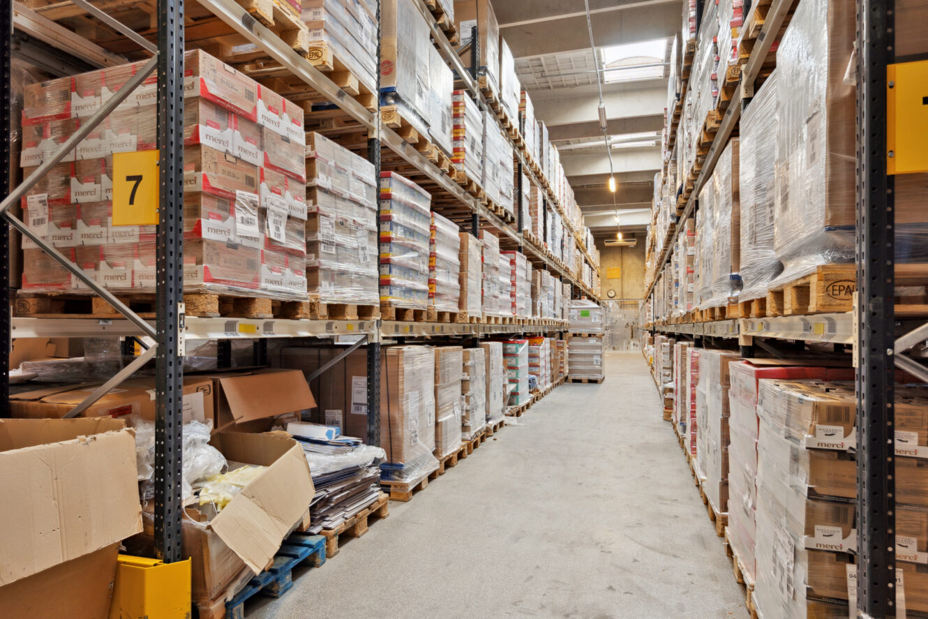 En lagergang med høje hylder stablet med paller med kasserede varer, nogle åbne papkasser på gulvet og lysstofrør over hovedet.